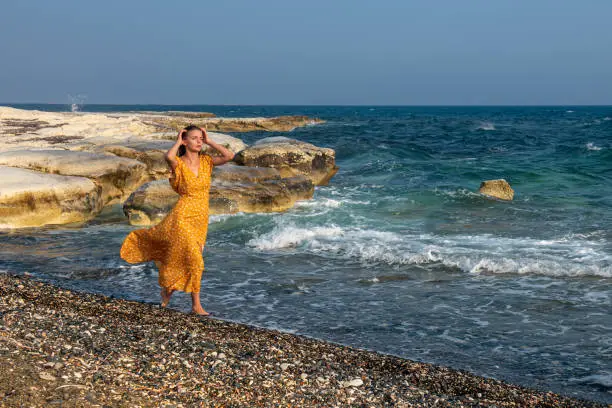 Young beautiful Caucasian woman wearing a yelllow summer dress and and walking on on the beach.