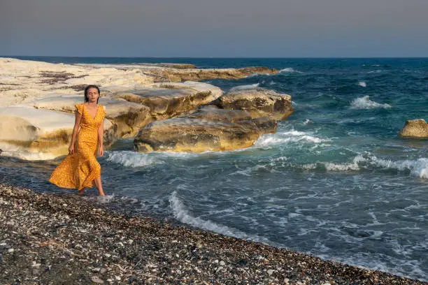 Young beautiful Caucasian woman wearing a yelllow summer dress and and walking on on the beach.