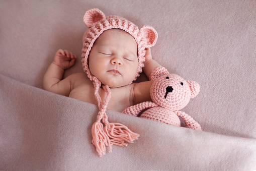 Portrait of cute newborn baby girl wearing pink hat with ears and sleeping with knitted teddy bear under pink blanket. Horizontal Image.