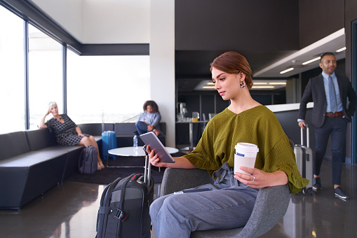 Fahionable millenial woman with brown hair holding coffee reading confidently on tablet, while waiting for flight in modern airport lounge during business trip