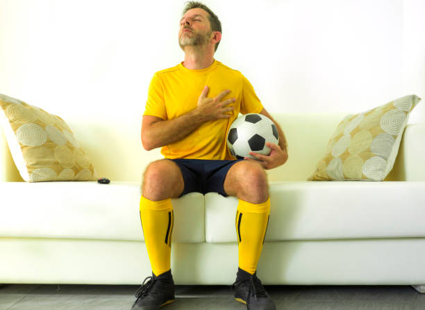 滑稽的肖像，歐洲足球隊制服的年輕人觀看足球比賽在電視在家庭沙發聽國歌與手在心臟在比賽期間熱情和情感 - indonesia football 個照片及圖片檔