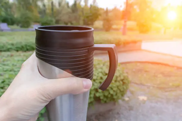 Season, autumn hot drinks and picnic concept - metal thermo coffee cup in woman hand sitting in park, coffee on the go.