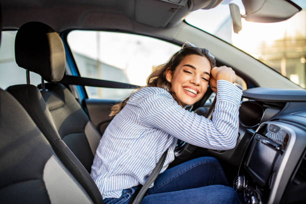 femme jeune et gai appréciant la nouvelle voiture étreignant le volant s'asseyant à l'intérieur - new automobile photos et images de collection