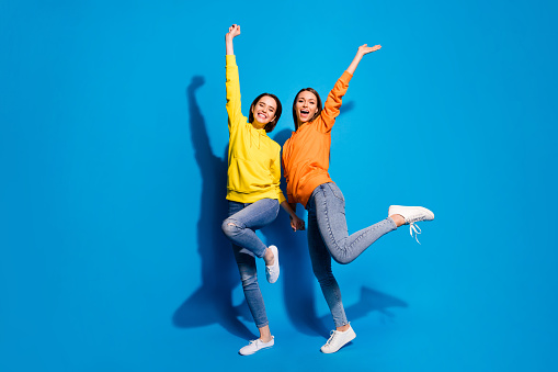 Full length photo of two pretty ladies raising arms up glad vacation walking traveling abroad wear casual bright hoodies and jeans isolated blue color background