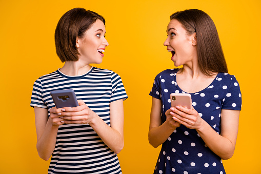 Photo of two pretty ladies holding telephones reading good news not believe luck wear summer, casual trendy t-shirts isolated yellow background