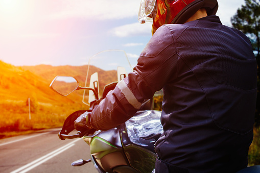 Close Up view of back of unrecognizable motor biker in helmet on motorcycle standing on edge of highway and looking on horizon with autumn  mountains at sunset.