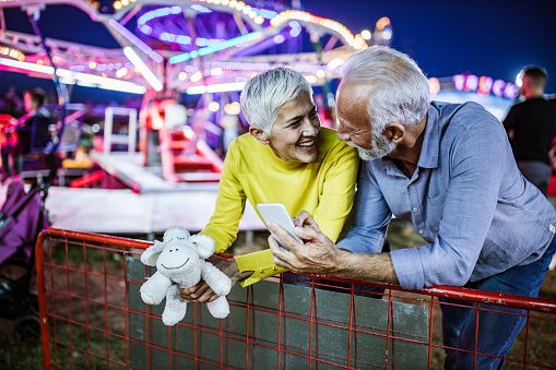 Happy senior couple communicating while using smart phone at amusement park by night.