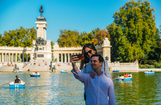 Madrid, Spain - Oct 5, 2019: Couple takes a selfie in front of boats across from monument to Alfonso XII in the Parque del Buen Retiro, known as the Park of the Pleasant Retreat in Madrid, Spain