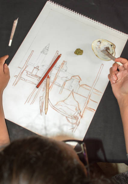istock Woman smoking a marijuana joint during a break after drawing. Background with cannabis bud, sketch and shell ashtray. Concept of drugs in the art. 1179337038