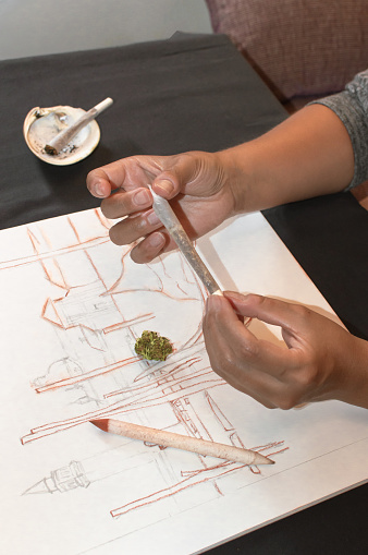 Artist rolling a joint of marijuana during a pause after drawing sketches. Background with cannabis bud, pencils and casual shell ashtray.