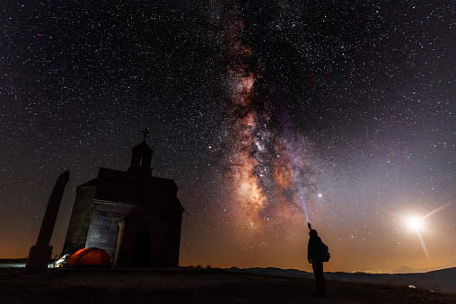Beautiful night landscape. Small chapel on the hill. Behind chapel bright milky way and moon. Person silhouette with flashlight illuminate starry sky.