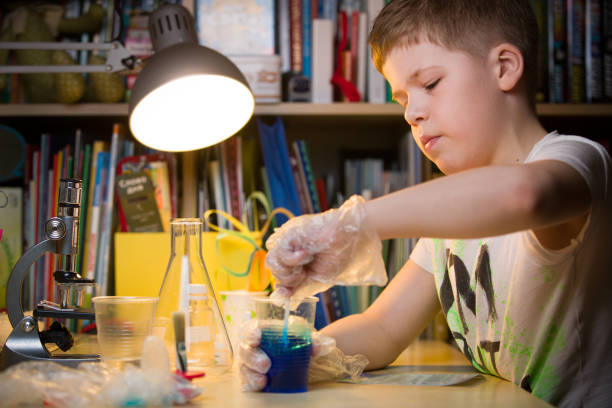 かわいい小学生の男の子が青い液体でグラスを持ってテーブルに座っています。ある若い科学者が自宅の研究室で実験を行う。屋内。子供と科学 - stem glass ストックフォトと画像