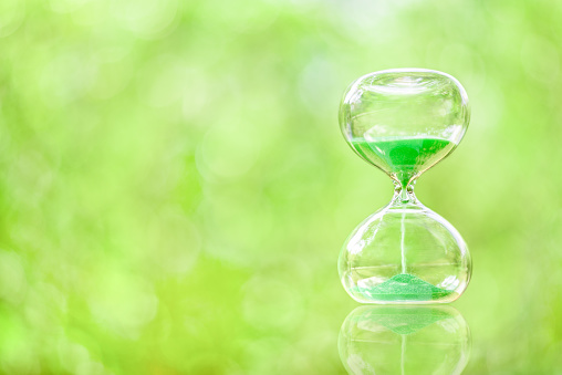 Time management, deadline and countdown concept : Green sand fall down in a clear glass sandclock or hourglass over green bokeh space, depicts flowing or passing of time from past, present to future