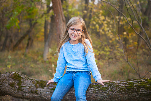 Portrait of a blonde girl with glasses outdoors in the park on the autumn park background sitting on the tree
