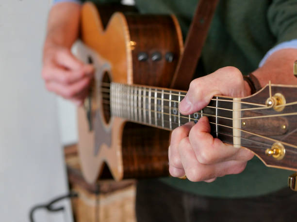 A man standing playing a guitar, with the focus on his left hand A man in a green woollen jumper standing playing the guitar. Focus on his left hand, with his fingers pressing down on the strings, while his right hand further down the guitar is out of focus as he strums the chords. He is wearing a brown leather strap to hold the wooden instrument securely while he plays his music. chord photos stock pictures, royalty-free photos & images