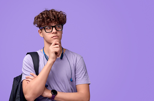 Charismatic teenager with backpack rubbing chin and looking away while thinking against violet background
