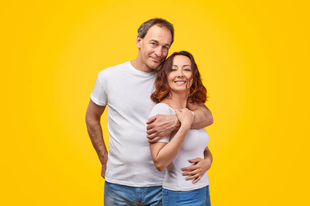 Happy mature couple looking at camera Adult man smiling and hugging cheerful woman while standing against bright yellow background together two men same clothes stock pictures, royalty-free photos & images