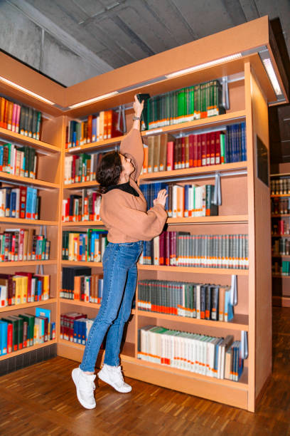 istock Young girl pulls book from top shelf of bookcase 1179418317