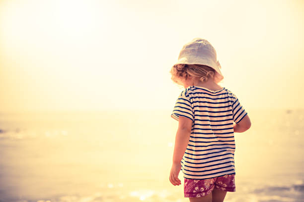 Child standing alone on beach with bright orange sunset sunlight during summer beach holidays concept for happy childhood travel lifestyle