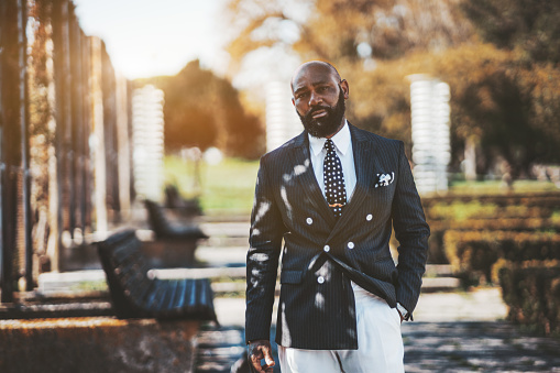 An elegant African man entrepreneur in a formal suit in a city park with benches in a defocused background; an adult fancy bald bearded black guy in a business formalwear on the street