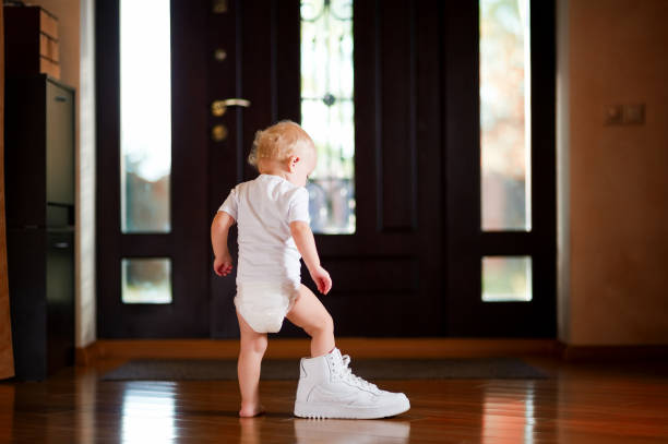 baby girl puts on a white dad's sneaker baby girl in a white T-shirt and diaper puts on a white dad's sneaker, standing in the hallway at home baby boutique stock pictures, royalty-free photos & images