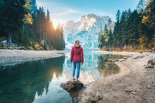 Woman is standing on the stone on Braies lake at sunrise in autumn. Dolomites, Italy. Landscape with girl, mountain, lake, beautiful reflection in water, trees, sky with sun. Forest in fall. Travel