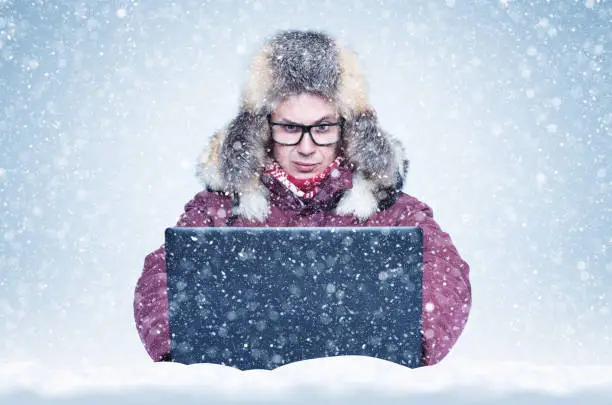 Photo of Frozen man in red winter clothes working on a laptop in the snow. Cold, frost, blizzard