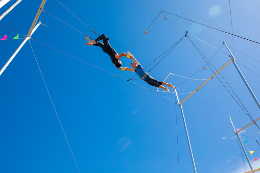 Two trapeze artists flying in the blue sky. One is jumping and the other is catching him.