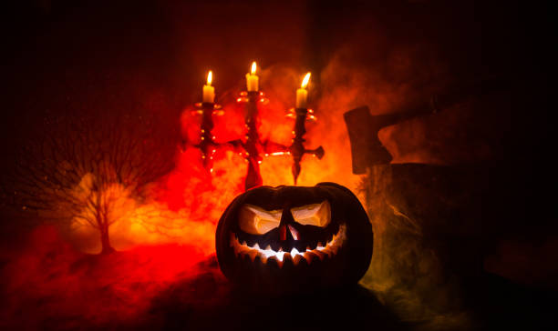 Scary orange pumpkin with carved eyes and a smile with burning candles and an ax Horror concept. Scary Halloween pumpkin with carved eyes and a smile with burning candles and an ax on a dark toned background. Empty space. Selective focus medieval restraints stock pictures, royalty-free photos & images