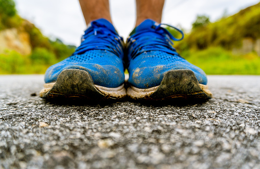 A close up shot of a worn pair of blue sneakers on tar road against a green and sky backdrop