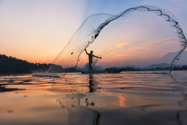 asiatische fischer werfen fischernetz in der dämmerung auf holzboot am see. konzept fisherman es lifestyle auf dem land. lopburi, thailand, asien - fishing net stock-fotos und bilder