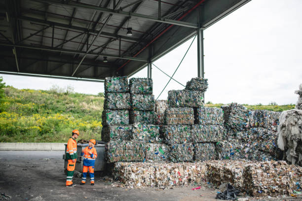 Coworkers Talking Next to Stacks of Compacted Recyclables Male and female waste management workers in protective suits and hardhats standing outdoors next to stacks of compacted and bundled recyclables. recycling center stock pictures, royalty-free photos & images