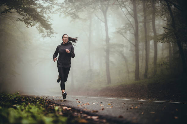 giovane donna atletica che fa jogging sulla strada nella foresta nebbiosa. - sport running exercising jogging foto e immagini stock