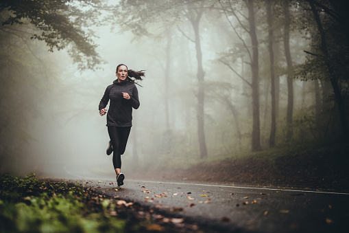 Young athletic woman jogging on the road in foggy forest.