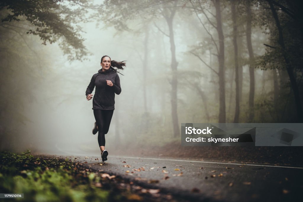 Young athletic woman jogging on the road in foggy forest. Determined athletic woman running through misty nature. Copy space. Running Stock Photo