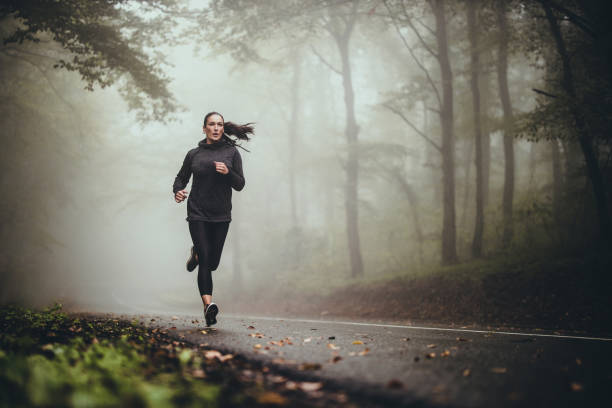 Young athletic woman jogging on the road in foggy forest. Determined athletic woman running through misty nature. Copy space. athleticism stock pictures, royalty-free photos & images