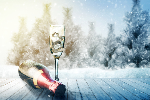 Champagne bottle and glass on wooden table with snowy fir trees background