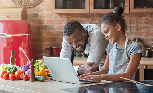 Smiling african daddy and daughter looking for nice recipe, using laptop at kitchen, empty space