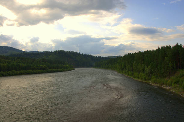 istock A wide, full-flowing river flowing through the forest under dramatic clouds illuminated by the rays of the setting sun. 1179525658