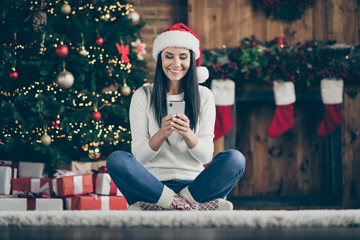 Full length photo of positive girl sir comfort floor carpet use her smartphone, type text messages search christmas discounts wear santa claus hat in house with x-mas decoration indoors