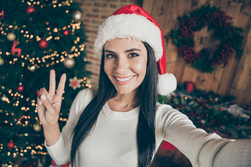 Enjoy christmas atmosphere. Close up photo of positive cheerful girl in santa claus, hat blogging on x-mas time take selfie make v-sign in room house with newyear wreath garlands decoration indoors