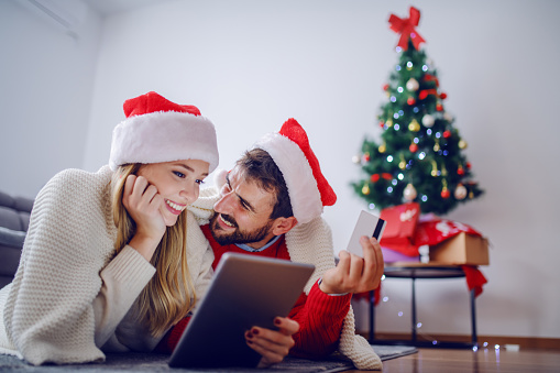 Cute Caucasian couple lying on stomach on the floor with santa hats on heads and using tablet for online shopping. Man holding credit card. Living room interior. In background is christmas tree.