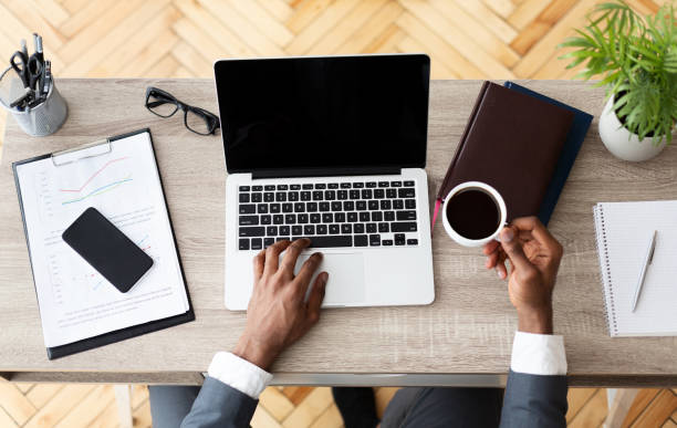 Office workplace top view. Black businessman working at wooden desk, using laptop and drinking coffee