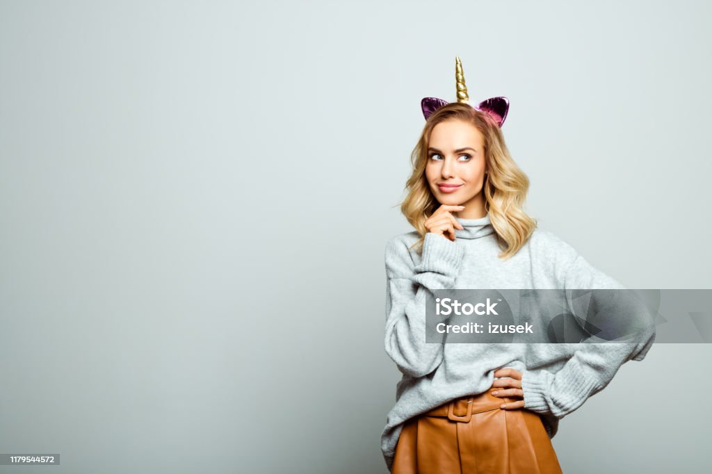 Studio portrait of beautiful woman with unicorn headband Studio shot of happy beautiful woman wearing sweater and unicorn headband standing against grey background and thinking with hand on chin. Unicorn Stock Photo