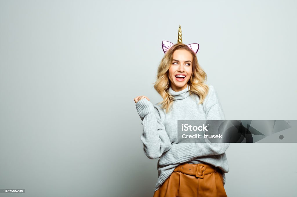 Studio portrait of surprised beautiful woman with unicorn headband Studio shot of happy beautiful woman wearing sweater and unicorn headband standing against grey background and laughing. 25-29 Years Stock Photo