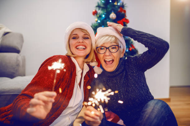 femme aînée heureuse et son descendant célébrant le nouvel an. les deux ayant des chapeaux de santa sur des têtes et retenant des cierges. à l'arrière-plan se trouve l'arbre de noel. concept de valeurs familiales. - senior adult photograph photography family tree photos et images de collection