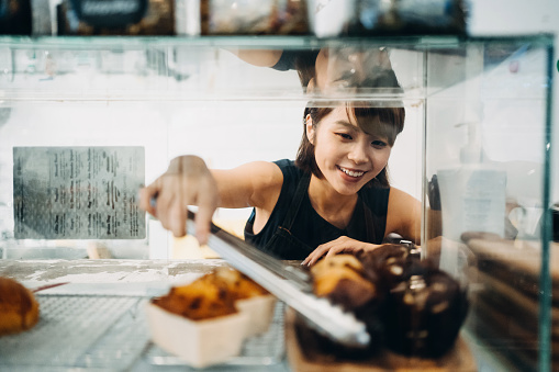 Cheerful young Asian woman working at a bakery and serving fresh bakery from the display cabinet