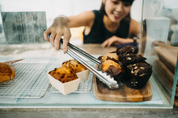 Cheerful young Asian woman working at a bakery and serving fresh bakery from the display cabinet