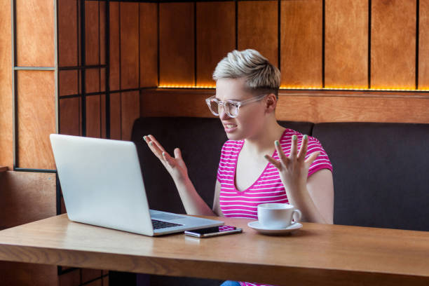 Young woman is sitting in cafe and having bad mood are admonishing a worker through a webcam with raised arms. Aggressive unhappy young girl freelancer with blonde short hair, in pink t-shirt and eyeglasses is sitting in cafe and having bad mood are admonishing a worker through a webcam with raised arms. clenching teeth stock pictures, royalty-free photos & images