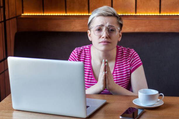 s'excuser! le portrait de jeune fille pigiste avec le cheveu court, dans le t-shirt et les lunettes roses s'assied dans le café et retenant la paume ensemble comme prient, regardant l'appareil-photo avec le visage heureux, - pleading women business sadness photos et images de collection